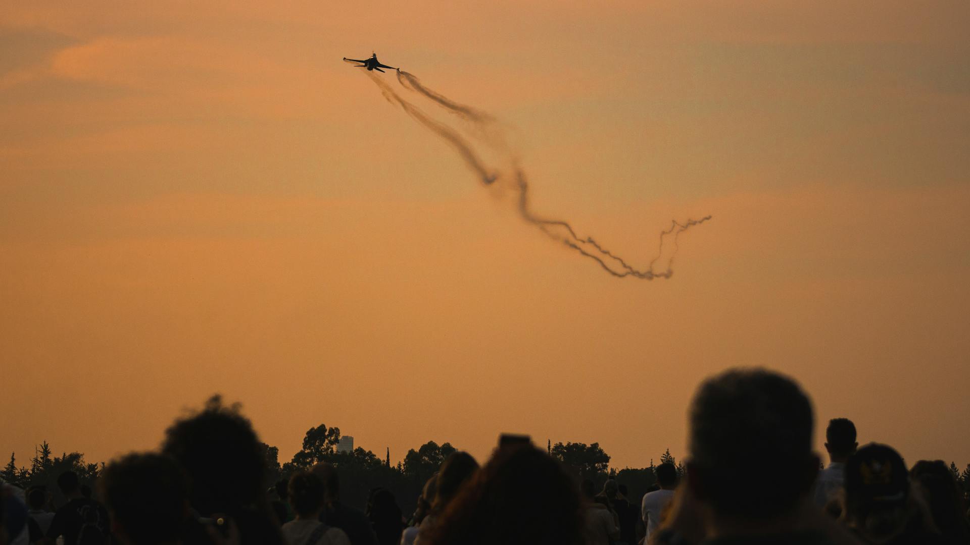 Jet flies over a crowd at airshow, leaving trails against a warm sunset sky.