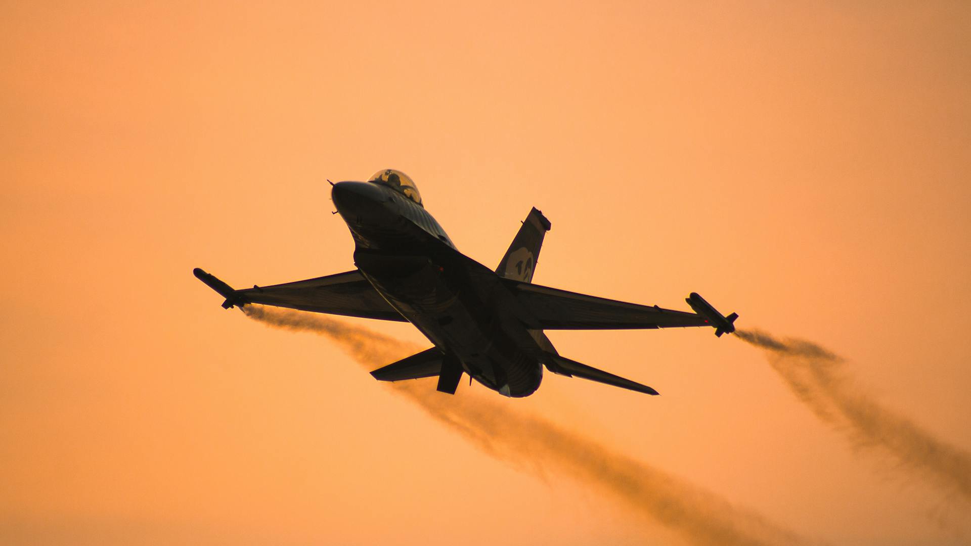 An F-16 jet flies silhouetted against an orange sunset sky, displaying a dramatic scene.