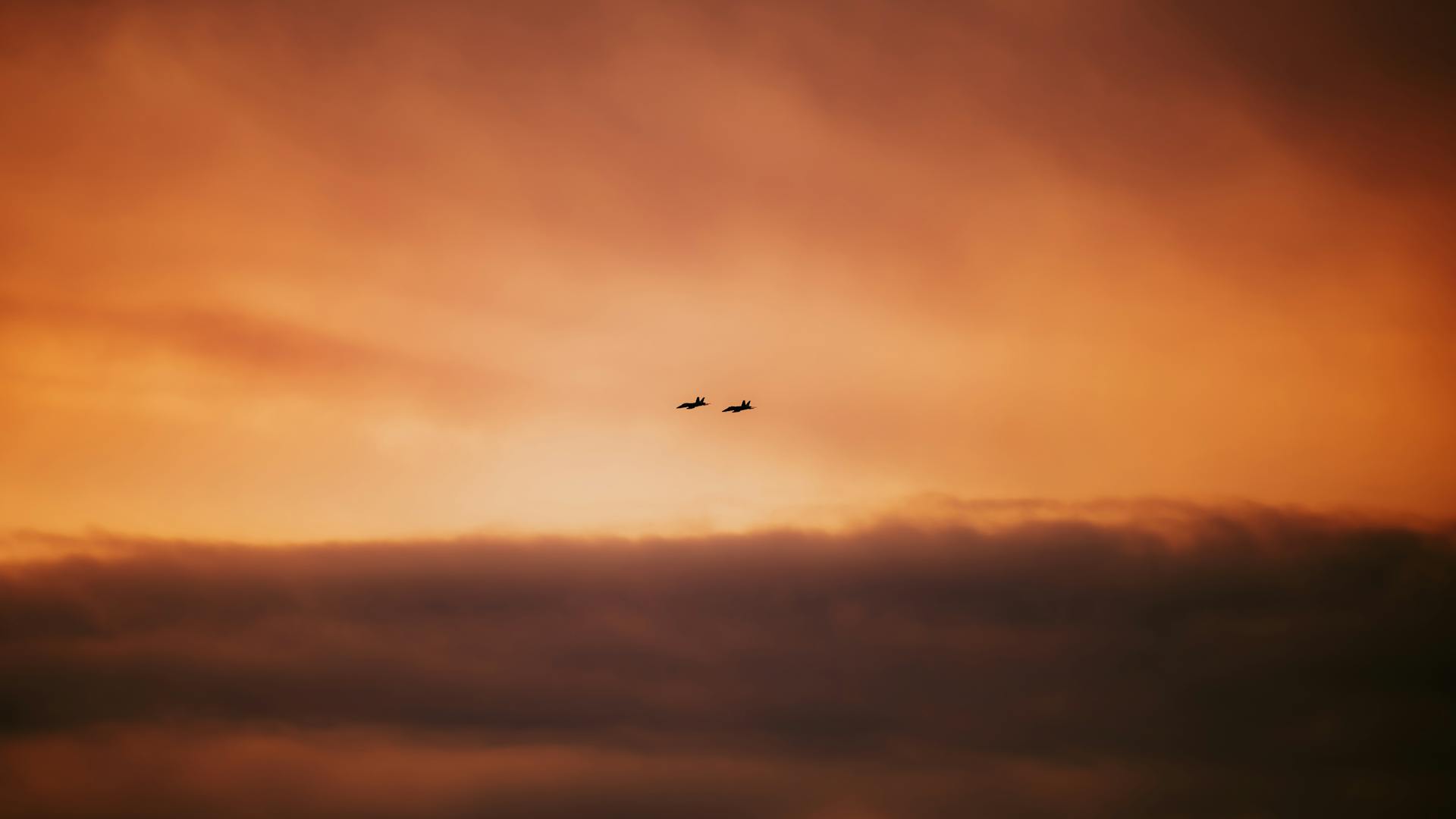 Two military fighter jets soar in silhouette against a vivid orange sunset sky, evoking a sense of power and tranquility.