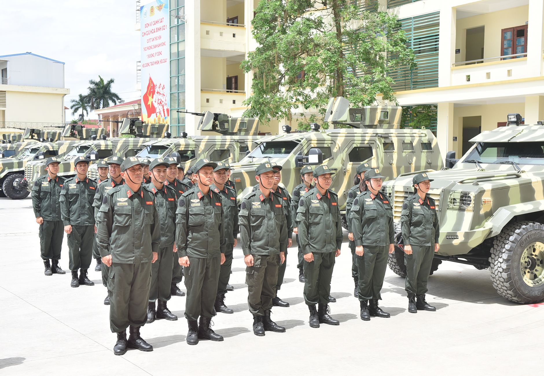 Group of military personnel in uniform stands in formation near armored vehicles on a sunny day.