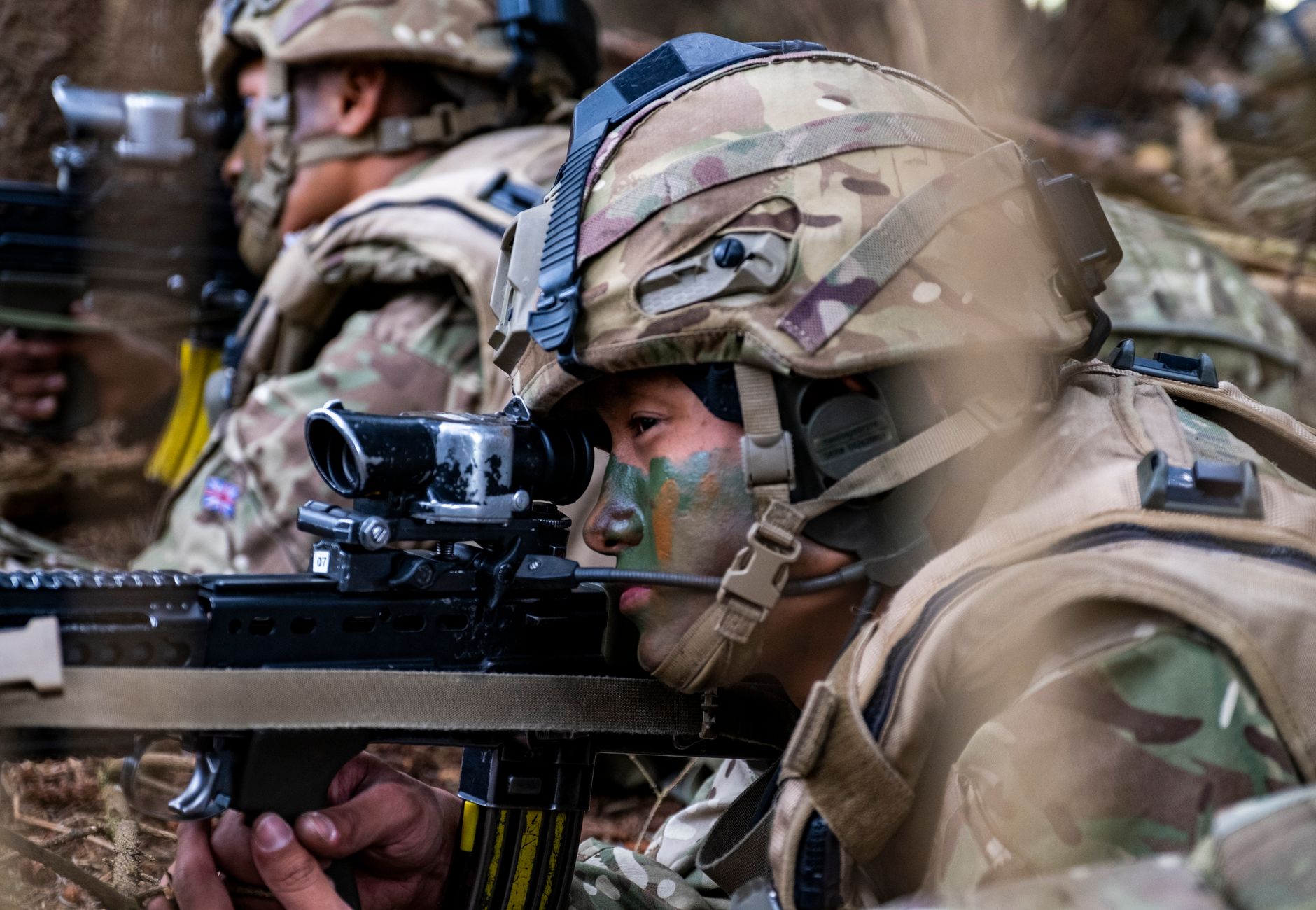Two soldiers wearing helmets and camouflage face paint taking aim during a military drill in a wooded area.